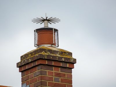 A red brick chimney stack fitted with a protective wire mesh bird guard and radiating metal anti-bird spikes, a preventative measure installed by professional chimney sweeps.