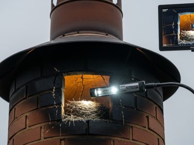 A gloved hand holding a specialized lighted camera to examine a bird's nest inside a brick chimney opening, with a connected monitor displaying the live feed during a CCTV chimney sweep inspection.