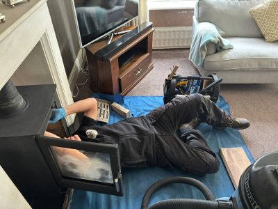 A professional technician lying on a blue protective sheet in a living room while reaching inside a wood-burning stove to perform routine chimney maintenance.